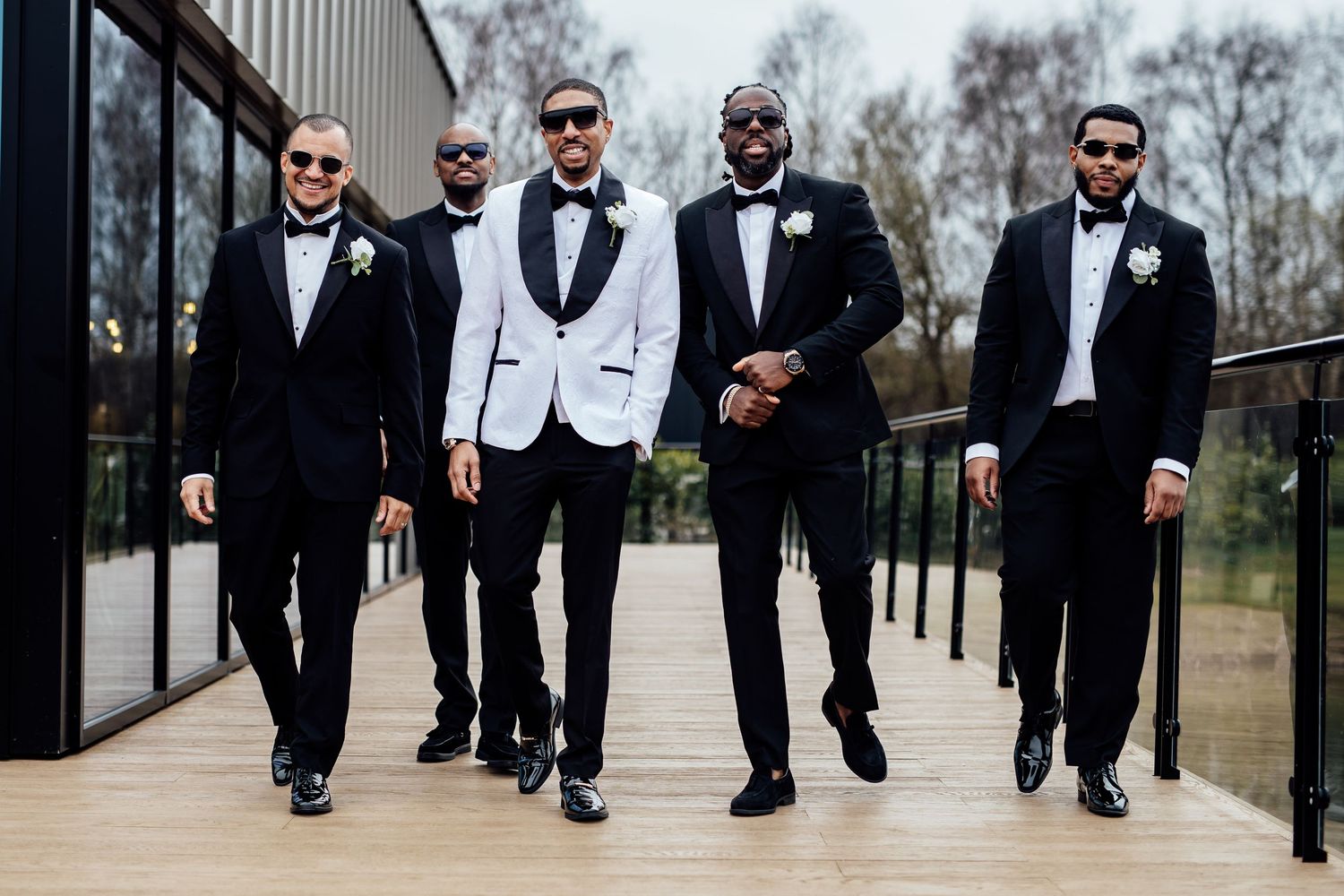 Groom in white tuxedo jacket poses with four groomsmen in black tuxedos on outdoor venue walkway.
