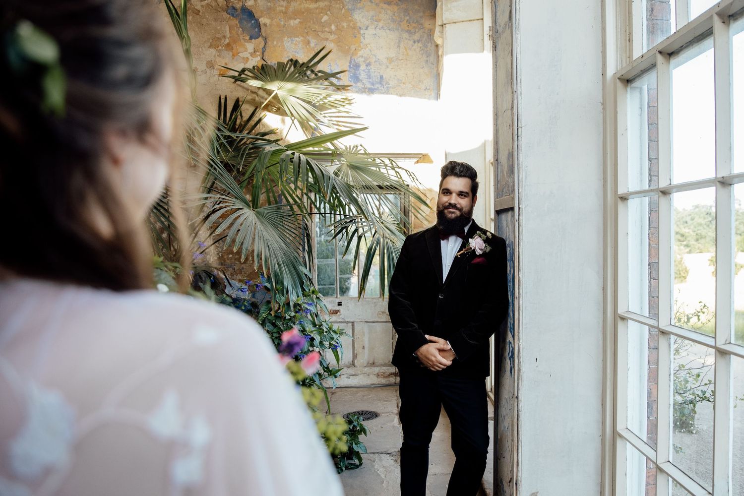A couple shares a first look moment in a bright sunlit room with tropical plants and French windows.