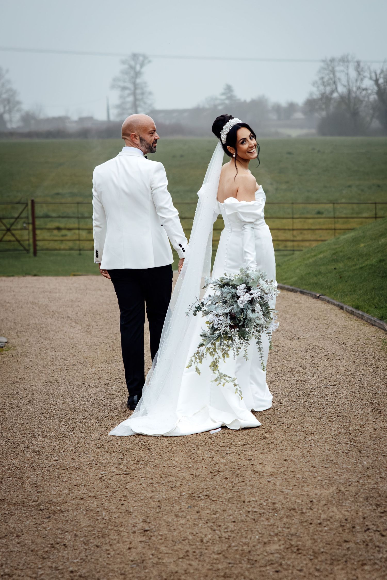 A couple in wedding attire walks together on a gravel path with misty fields and trees in the background.