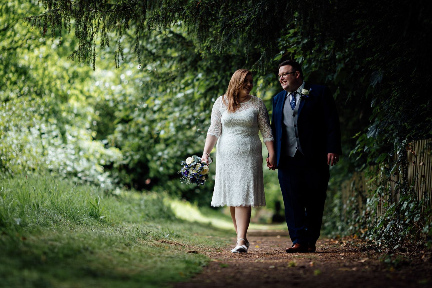 Couple walking along a tree-lined path in a lush green garden setting.