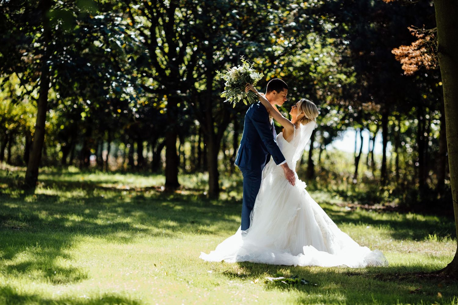 A couple embracing in a scenic wooded area with sunlight filtering through trees.