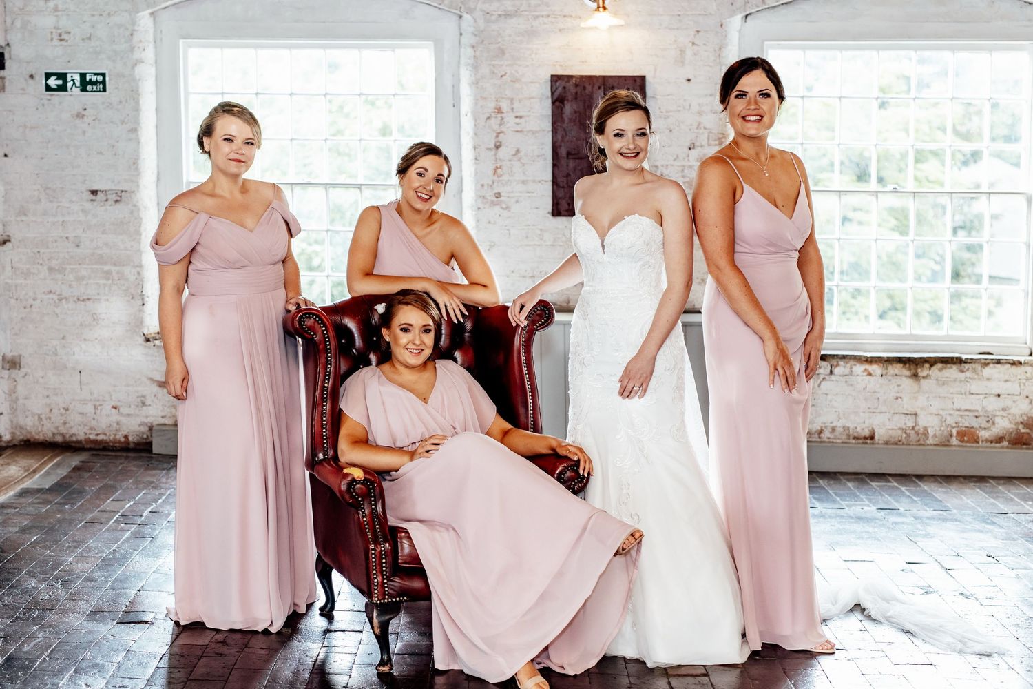 Bridesmaids in blush pink dresses gather in an industrial-style venue with exposed brick walls.