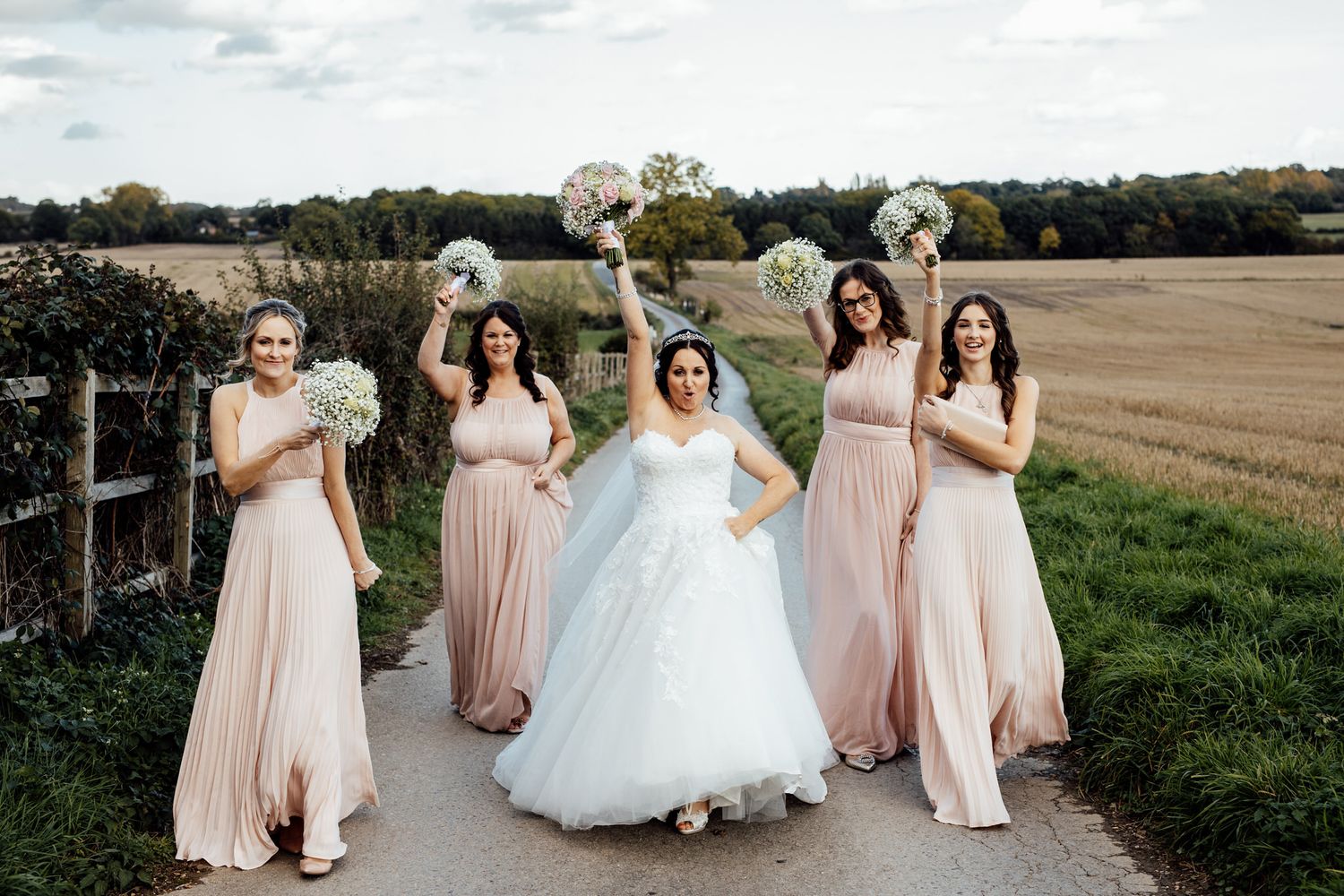 Bridal party in pink and white dresses walking through a scenic countryside field.