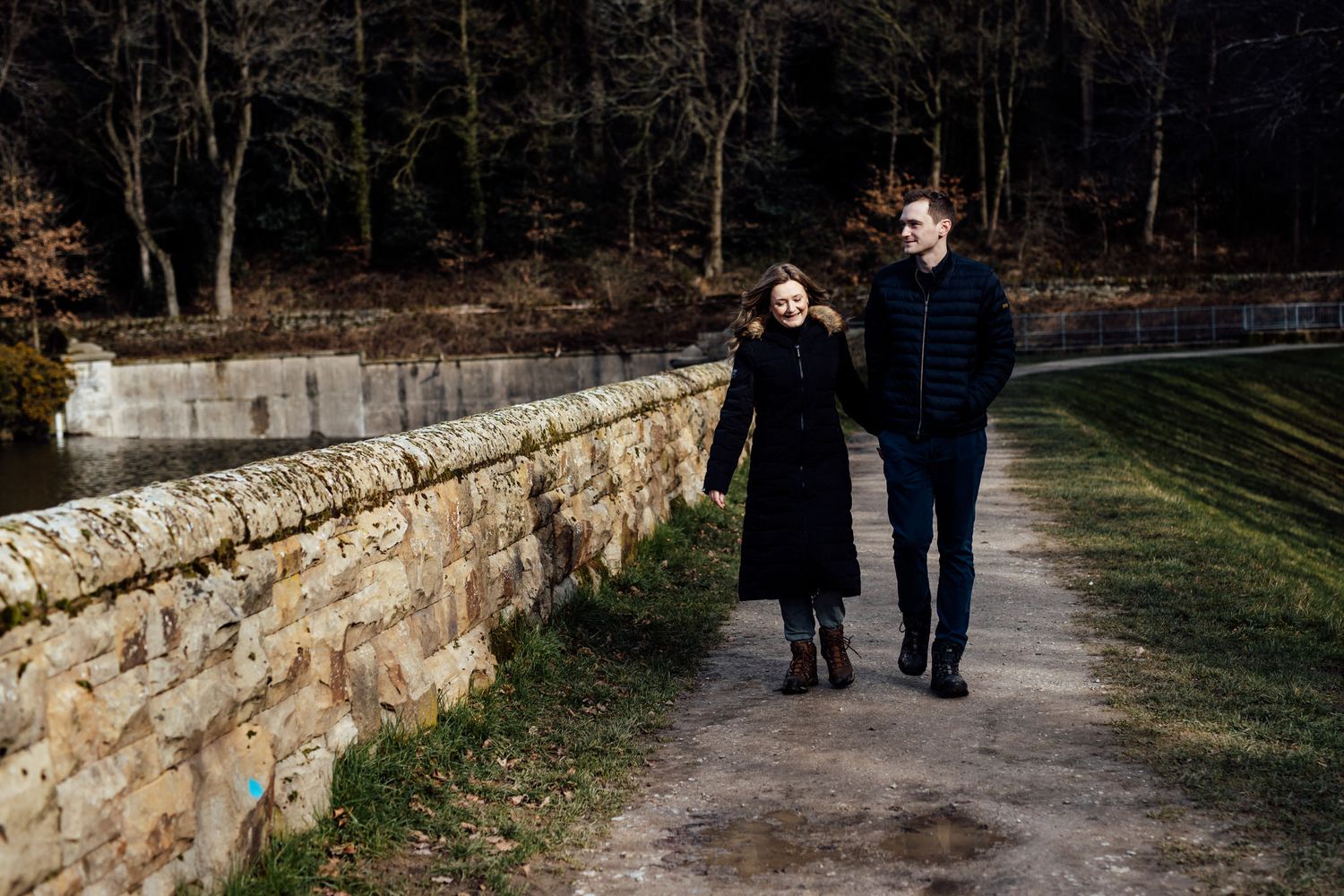 A romantic couple strolls along a stone wall path surrounded by bare winter trees.