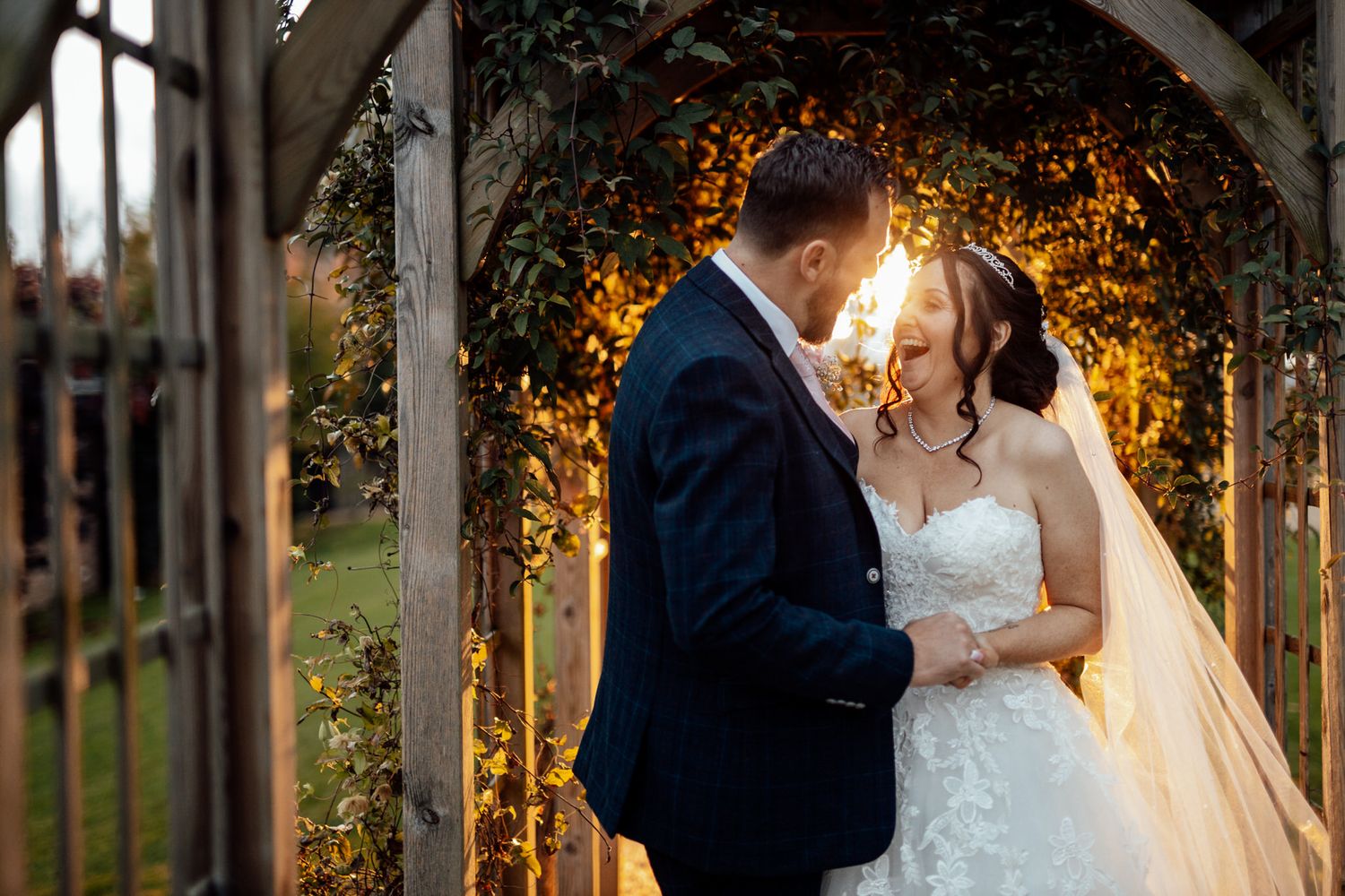 Romantic outdoor wedding portraits with couple framed by illuminated garden arbor at sunset.
