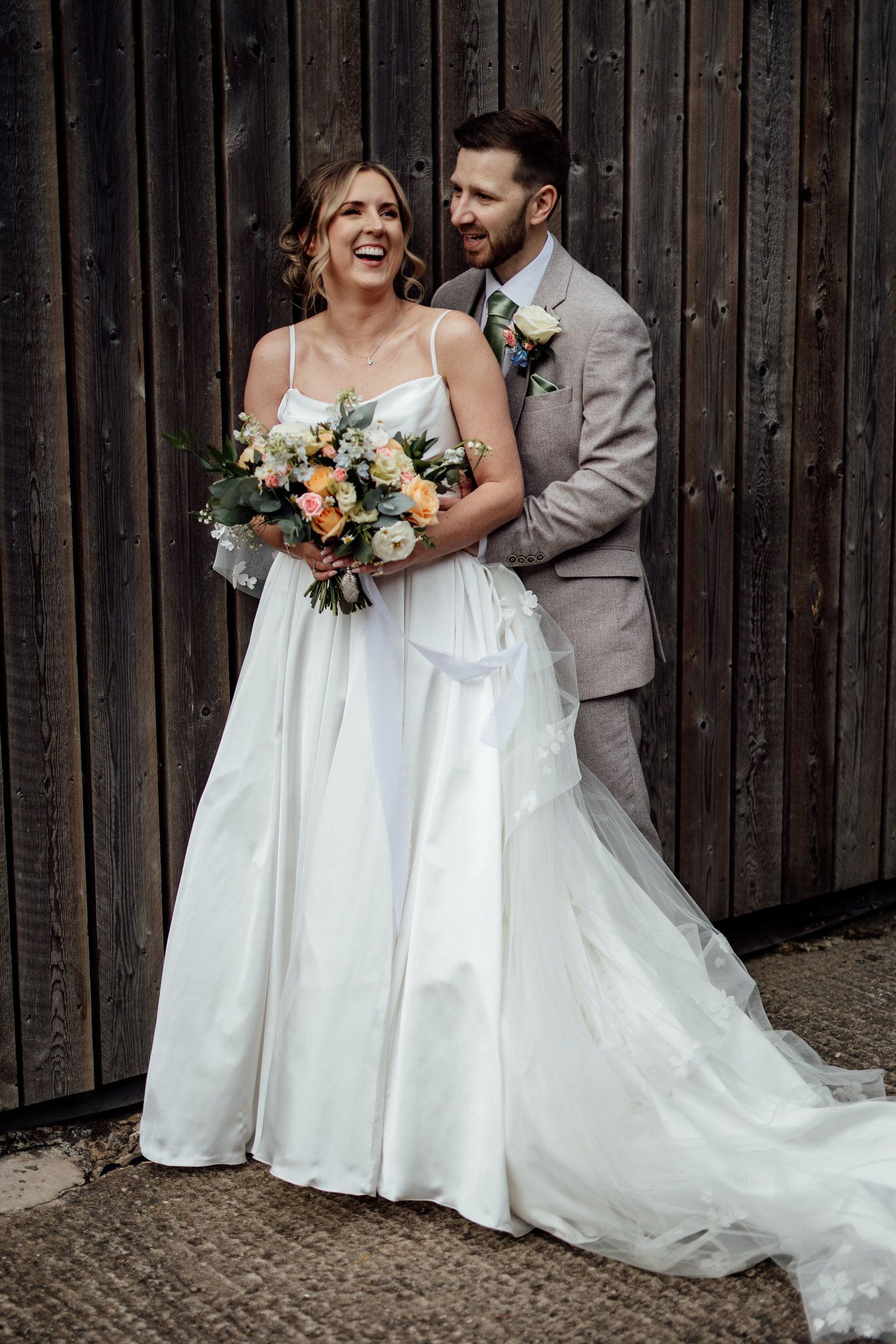 A joyful wedding portrait against rustic wooden walls with a beautiful floral bouquet.