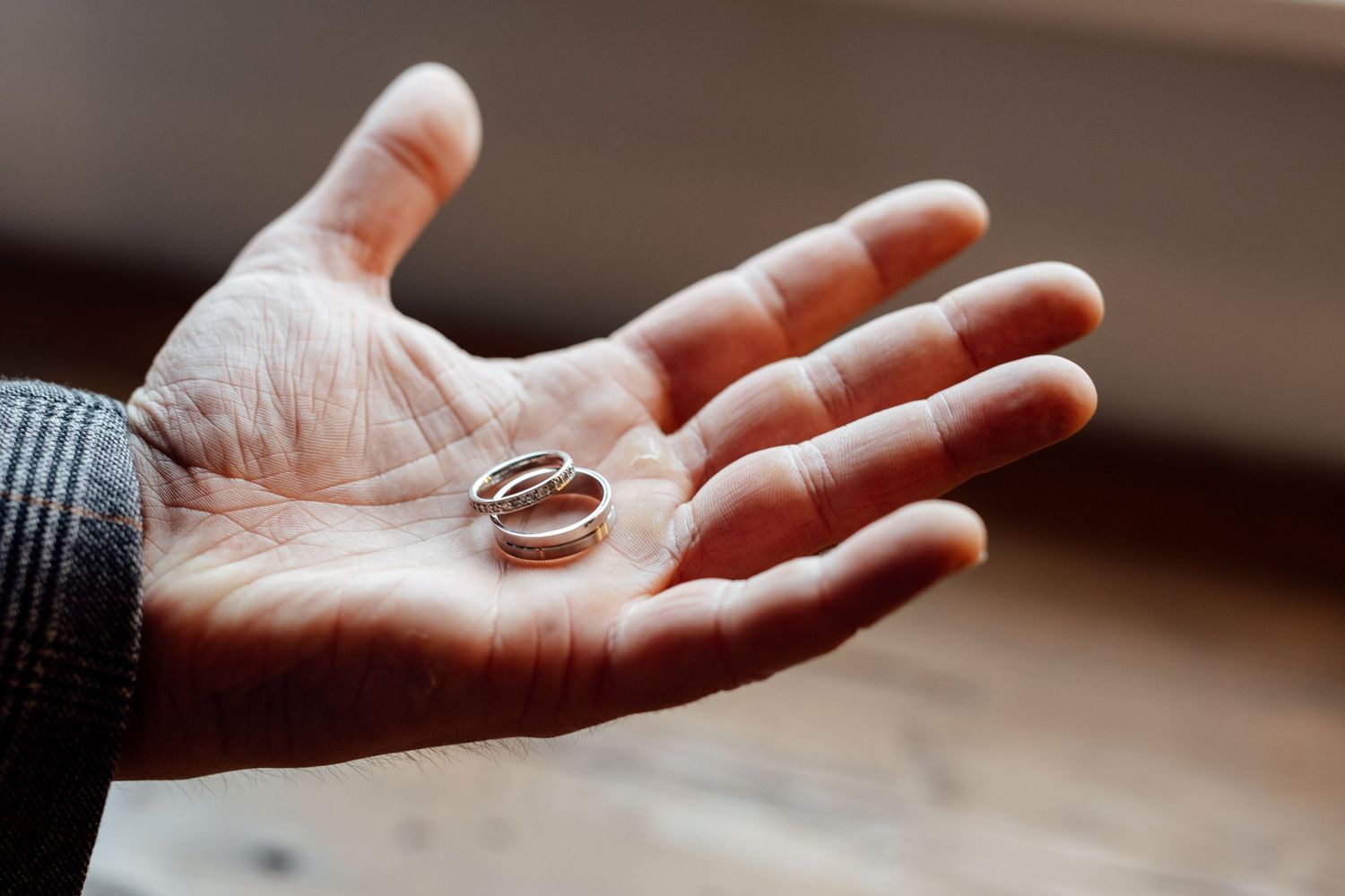 Wedding rings rest in an open palm wearing a gray sleeve against a blurred background.