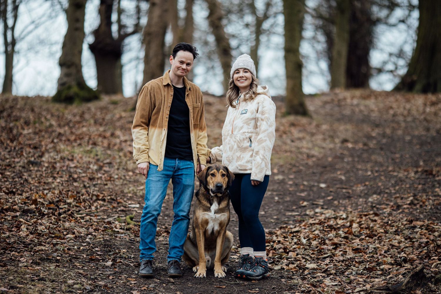 A couple standing with their dog on a forest trail covered in fallen leaves.