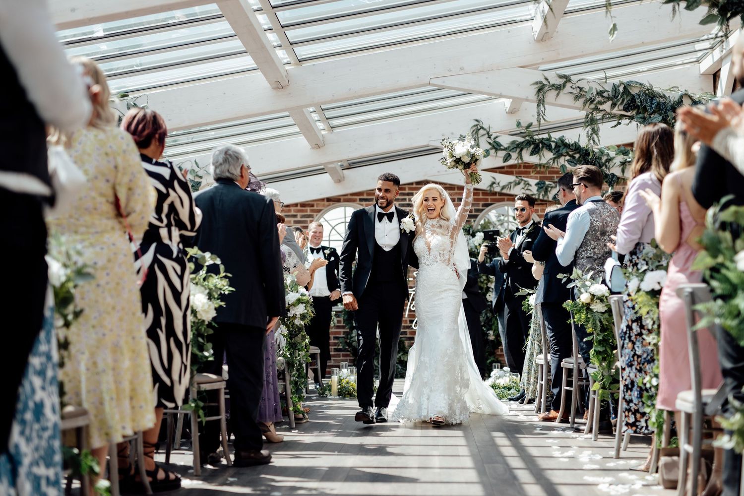 Wedding ceremony in progress under a light-filled conservatory with floral decorations.