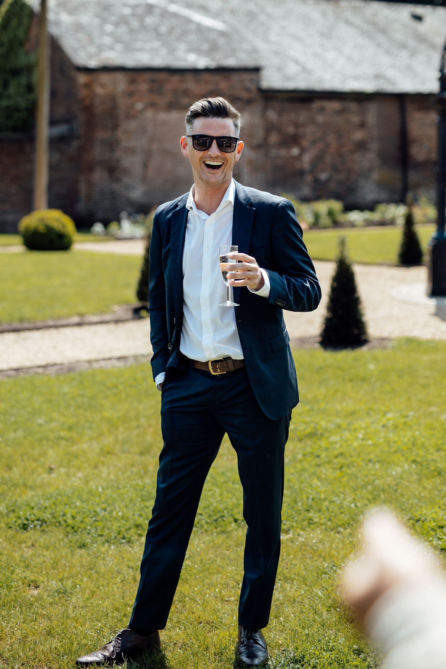 A person in a navy suit and sunglasses poses playfully on a grassy lawn near brick buildings.
