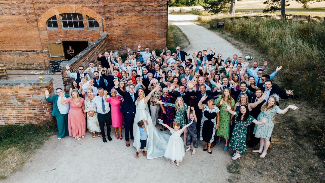 Aerial view of large wedding group photo outside historic brick building near water.