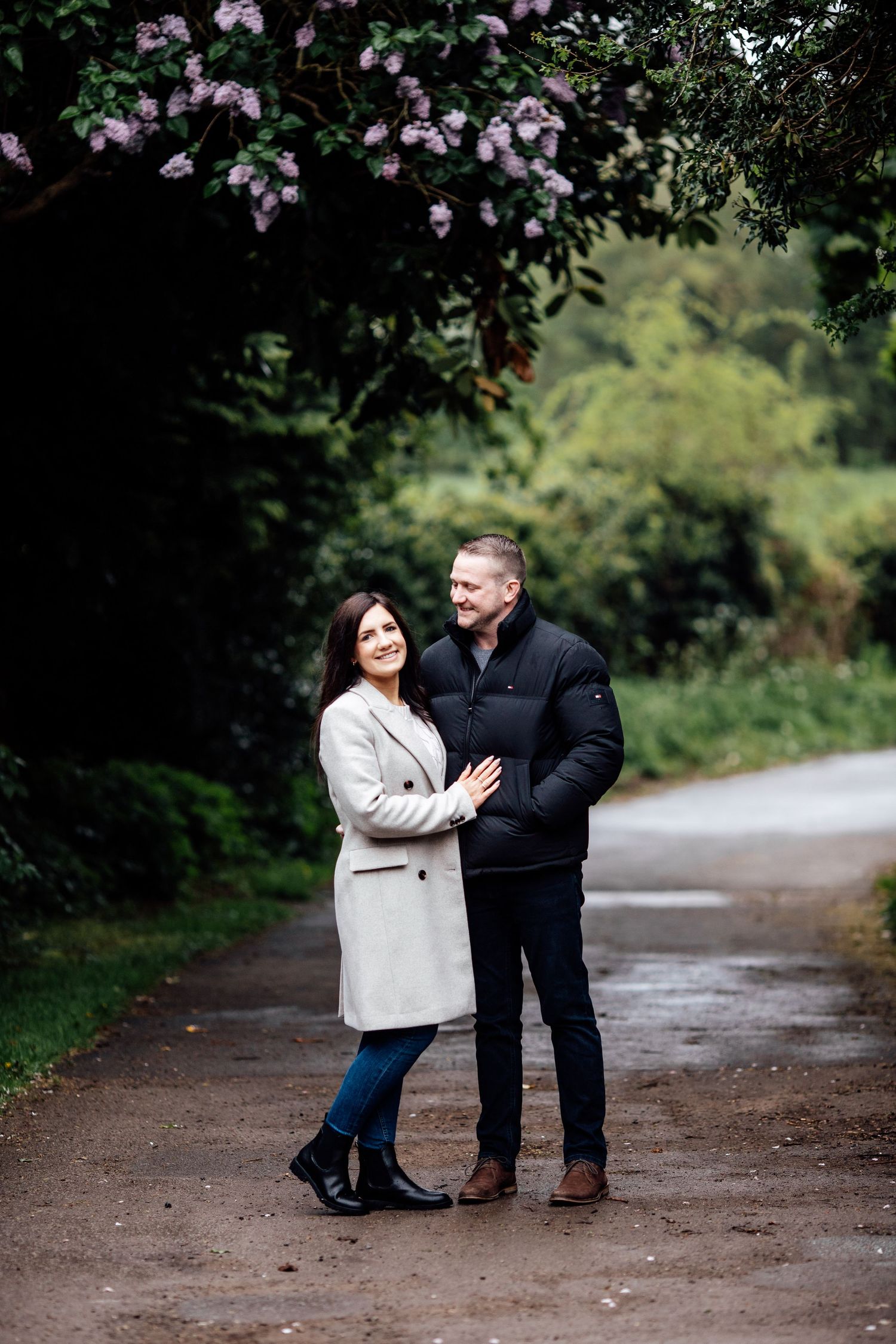 A couple shares a tender moment on a dirt path surrounded by blooming pink flowers and greenery.