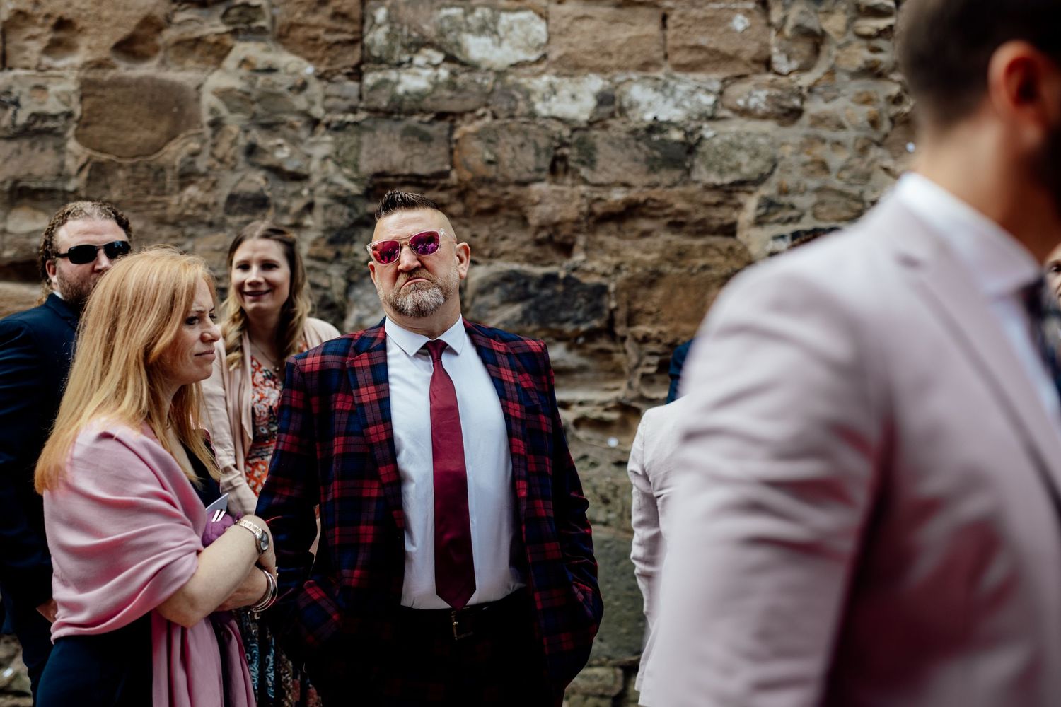 Wedding guests socializing and laughing at an outdoor reception by a stone wall.