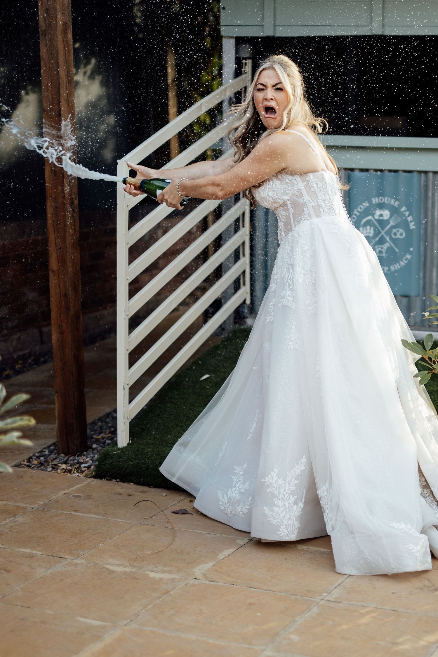 Bride in white gown dramatically popping champagne bottle at outdoor wedding venue.