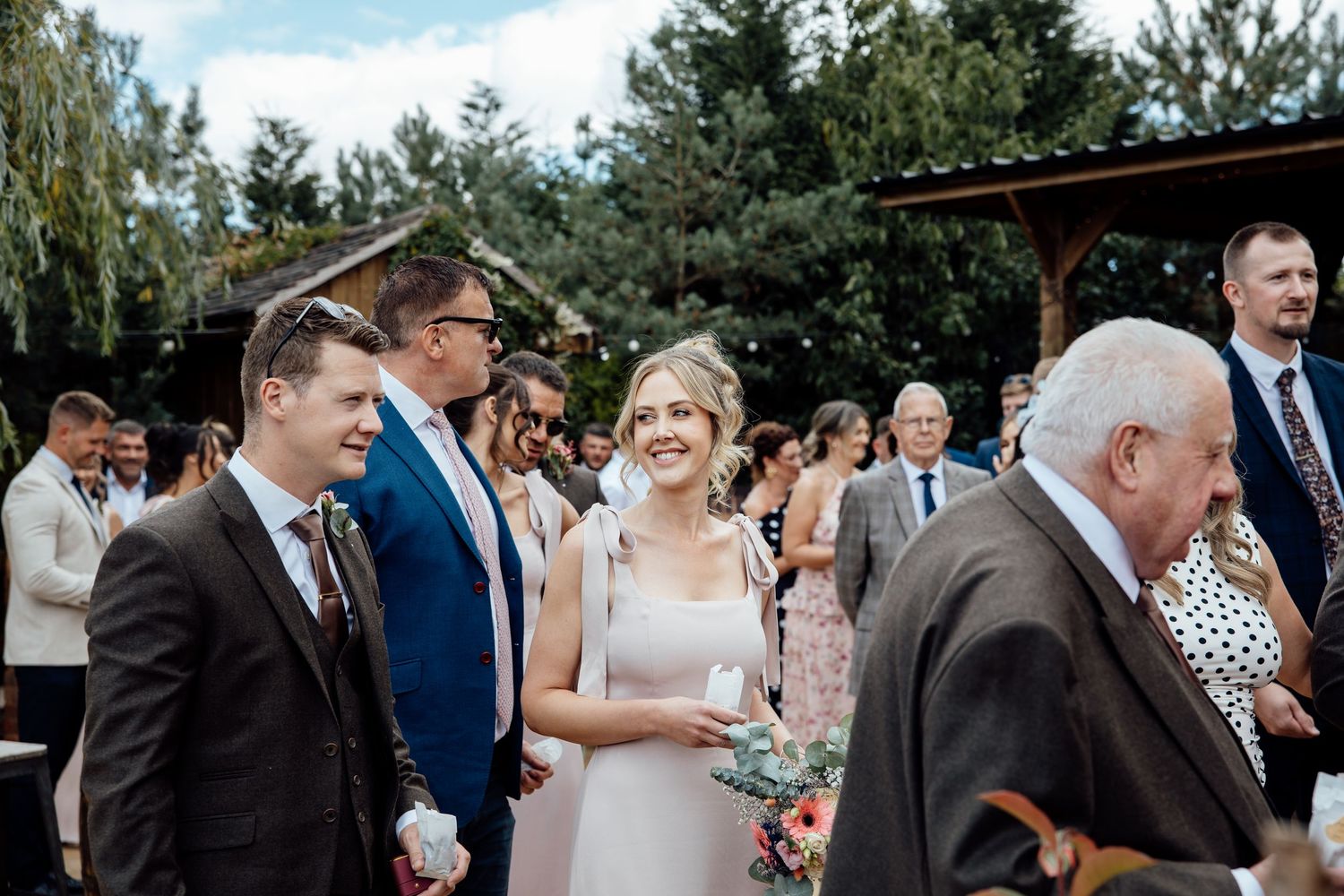 Bride smiling at outdoor wedding ceremony surrounded by guests in elegant attire.