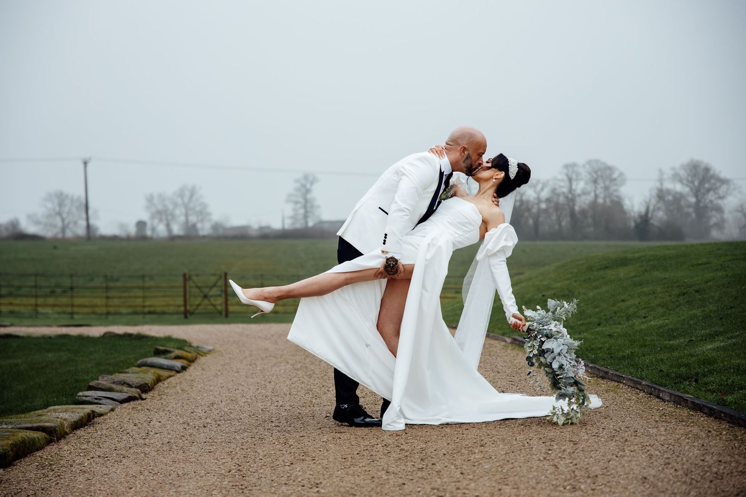 Wedding couple performing romantic dip poses on an outdoor path surrounded by green fields.