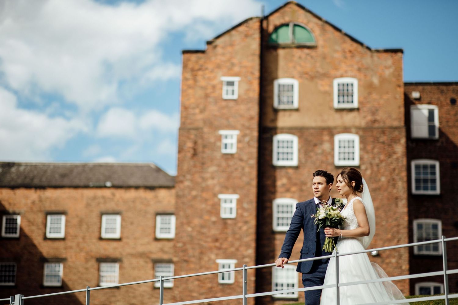 Bride and groom pose outside historic brick building under blue sky.