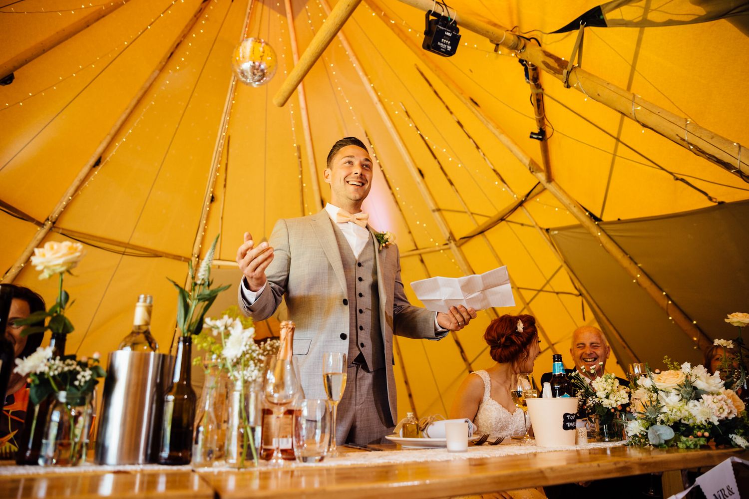 Wedding reception setup inside a warmly lit tipi tent with hanging decorations and table settings.