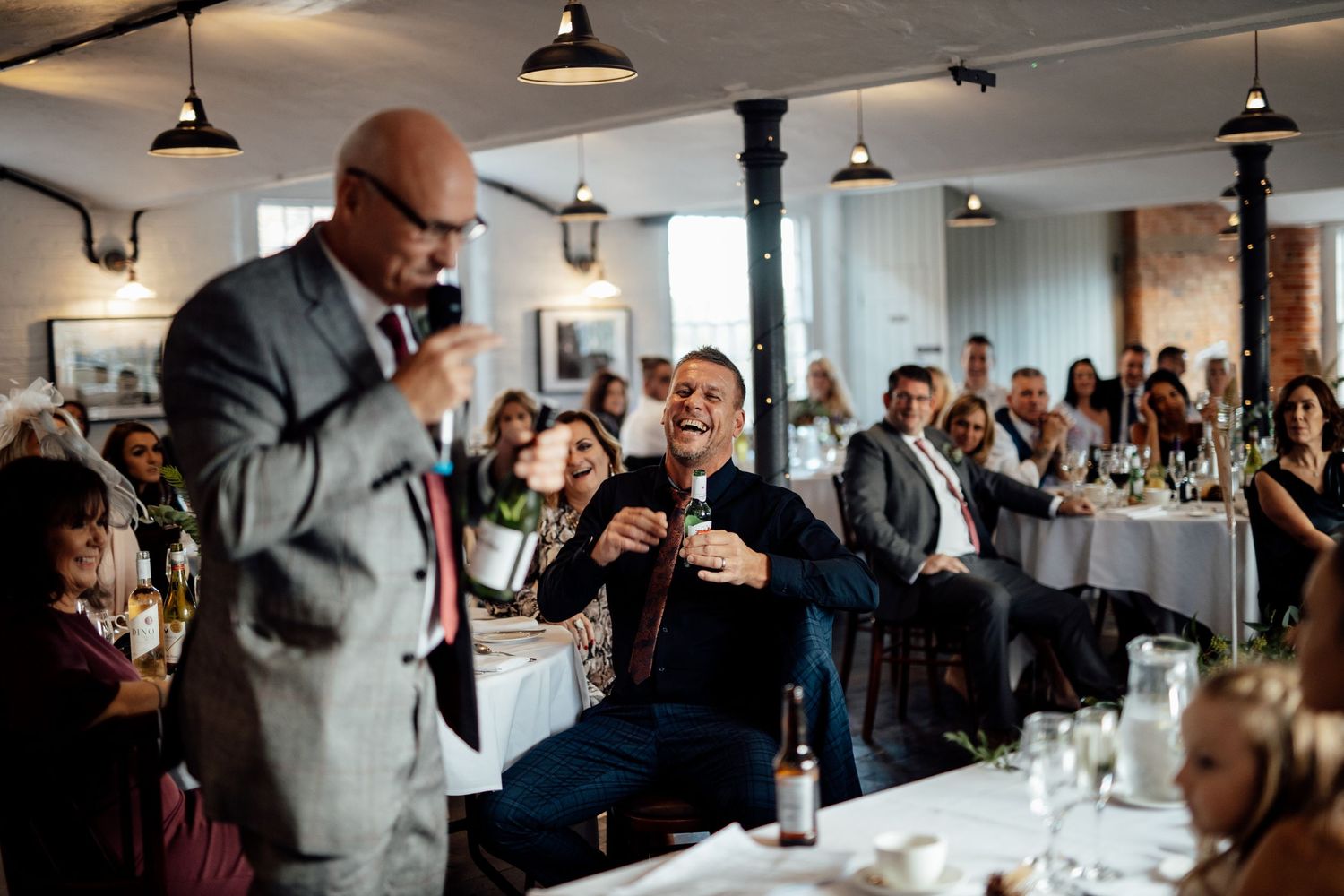 Wedding guests laugh and celebrate during a reception speech in a rustic venue with exposed brick walls and pendant lighting.