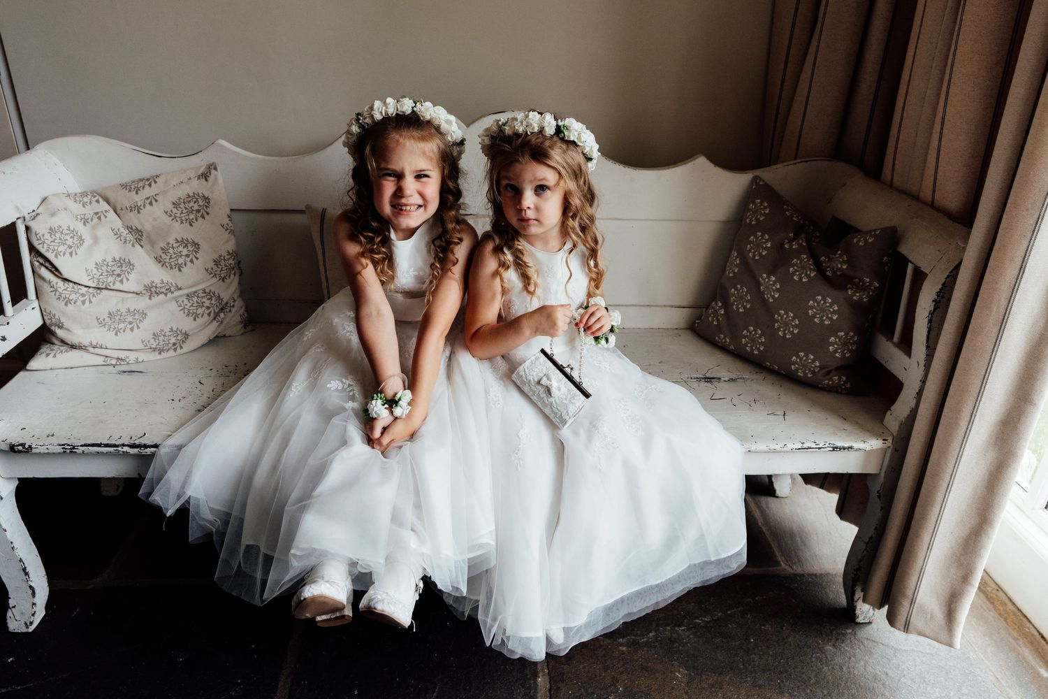 Two flower girls in white tulle dresses sitting together on an upholstered bench.