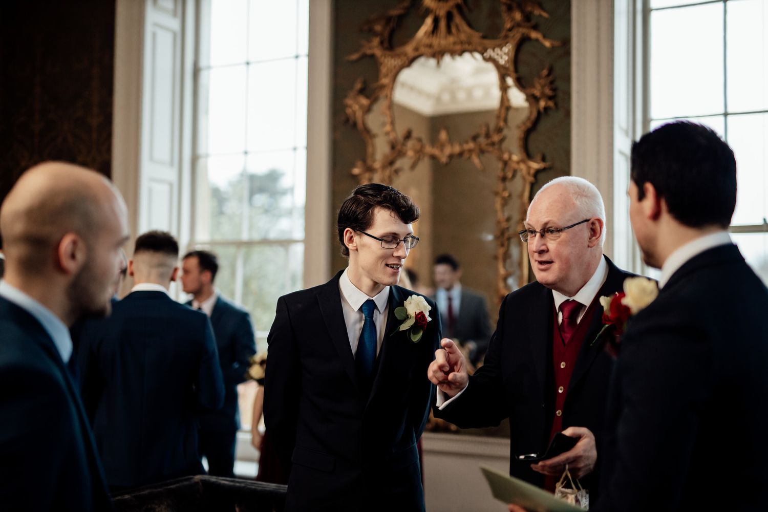 Wedding guests mingling in an elegant room with tall windows and ornate moldings.