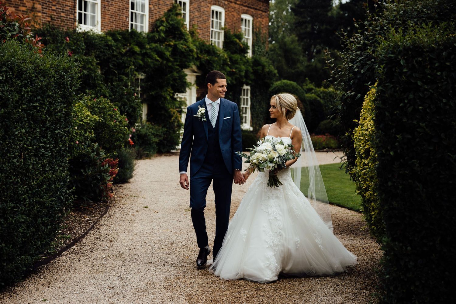 A couple walking along a garden path beside a historic brick building.