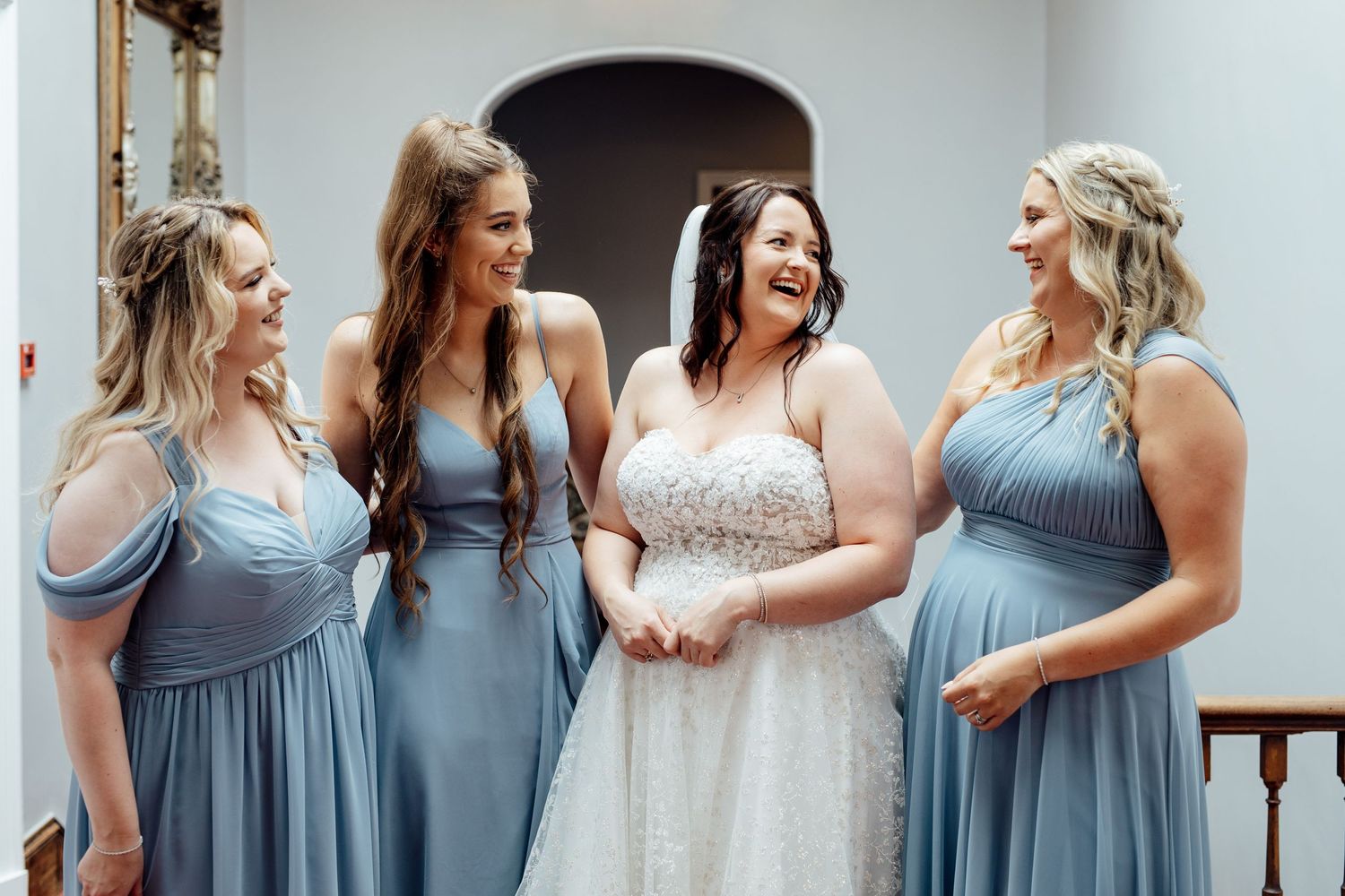 Wedding party in matching blue gowns and white dress share joyful laughter indoors.