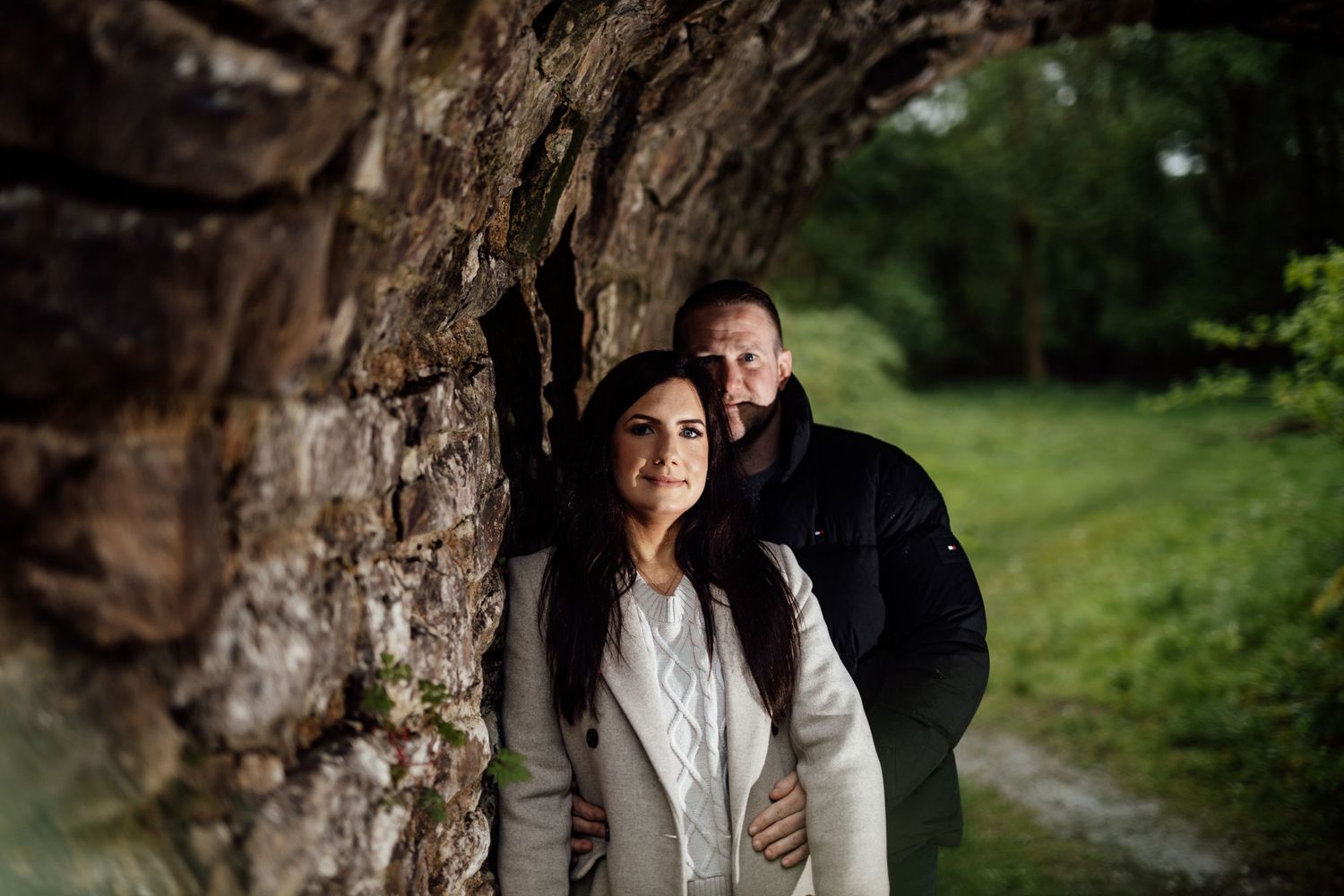 A romantic couple shares an intimate moment under a stone archway in a lush green park setting.