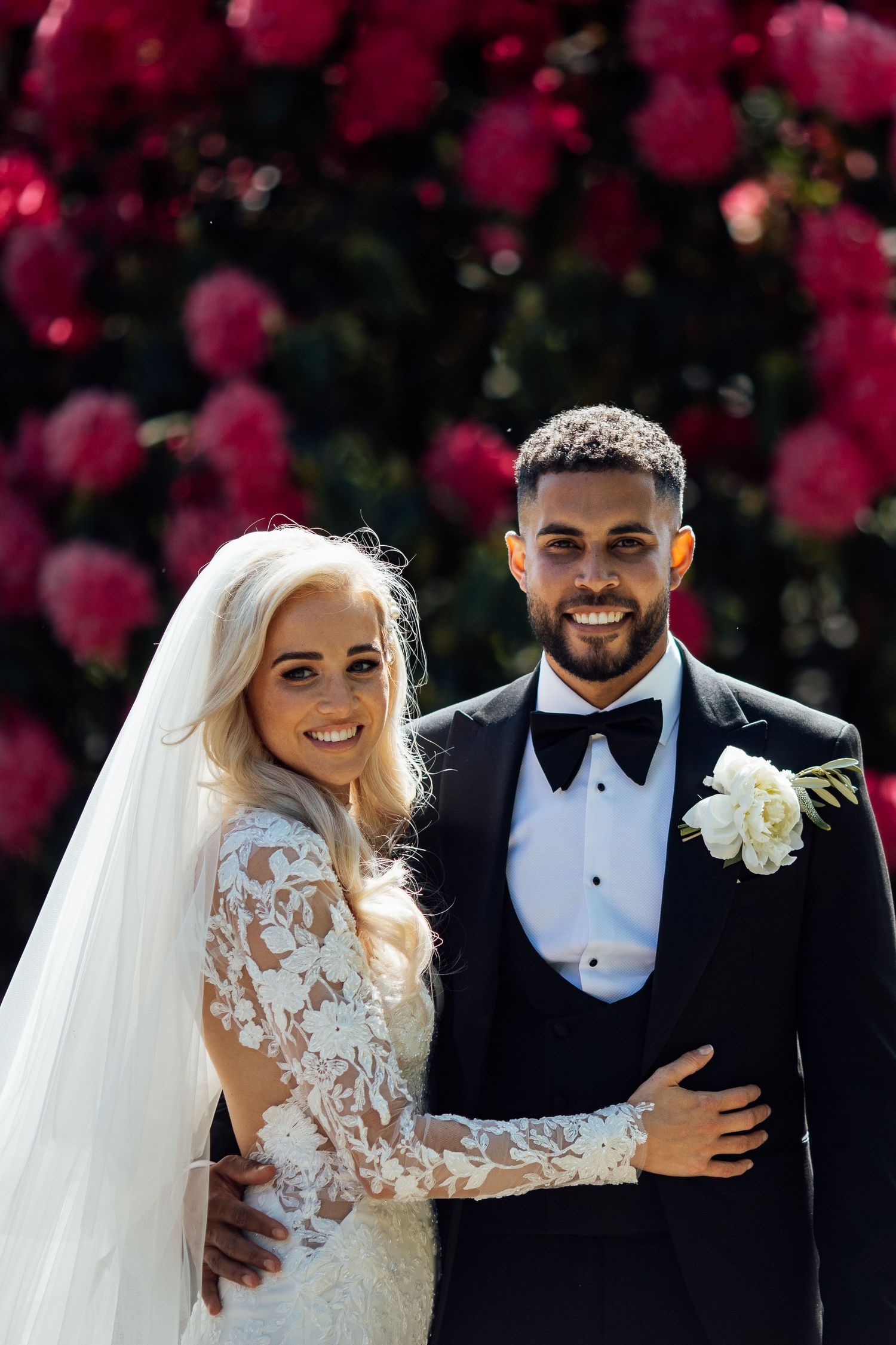 Couple in wedding attire poses in front of vibrant pink flowering trees.