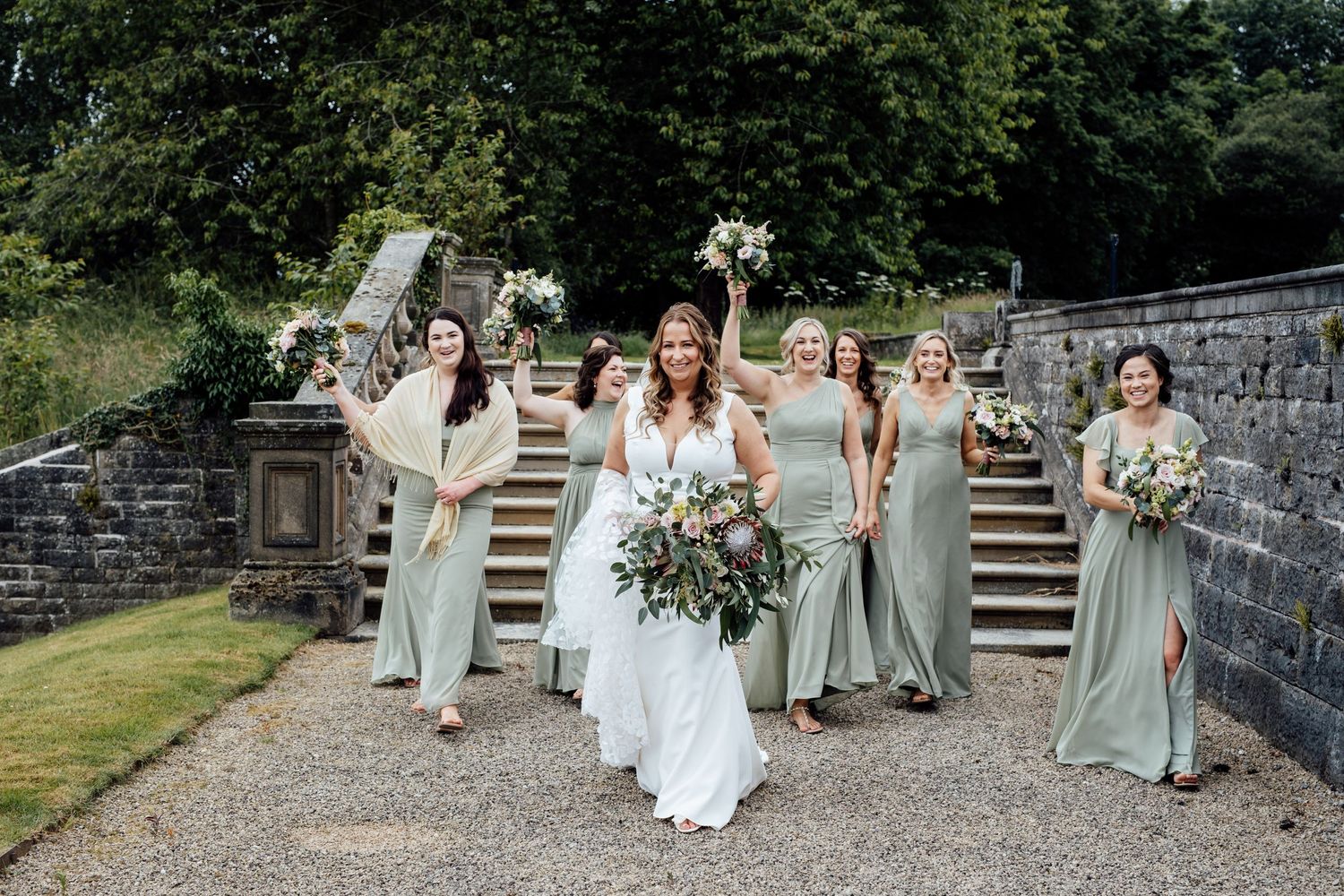 Wedding party in sage green dresses poses on stone steps outdoors.