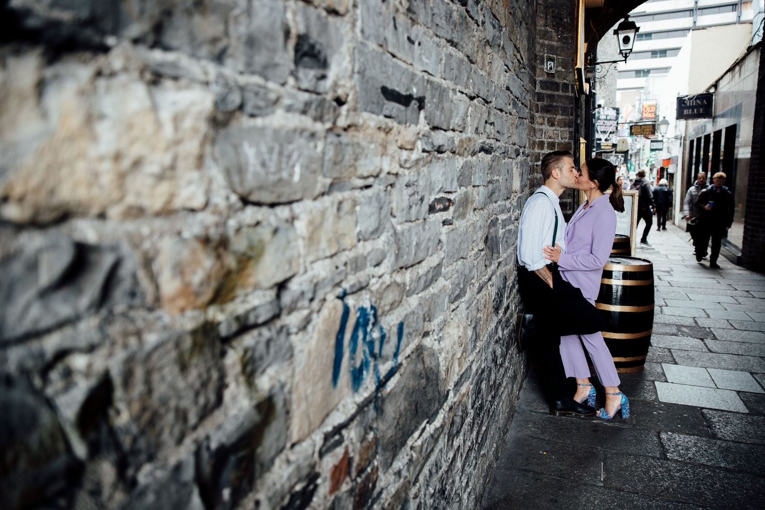 Couple sits against stone wall on urban street with pedestrians walking by.
