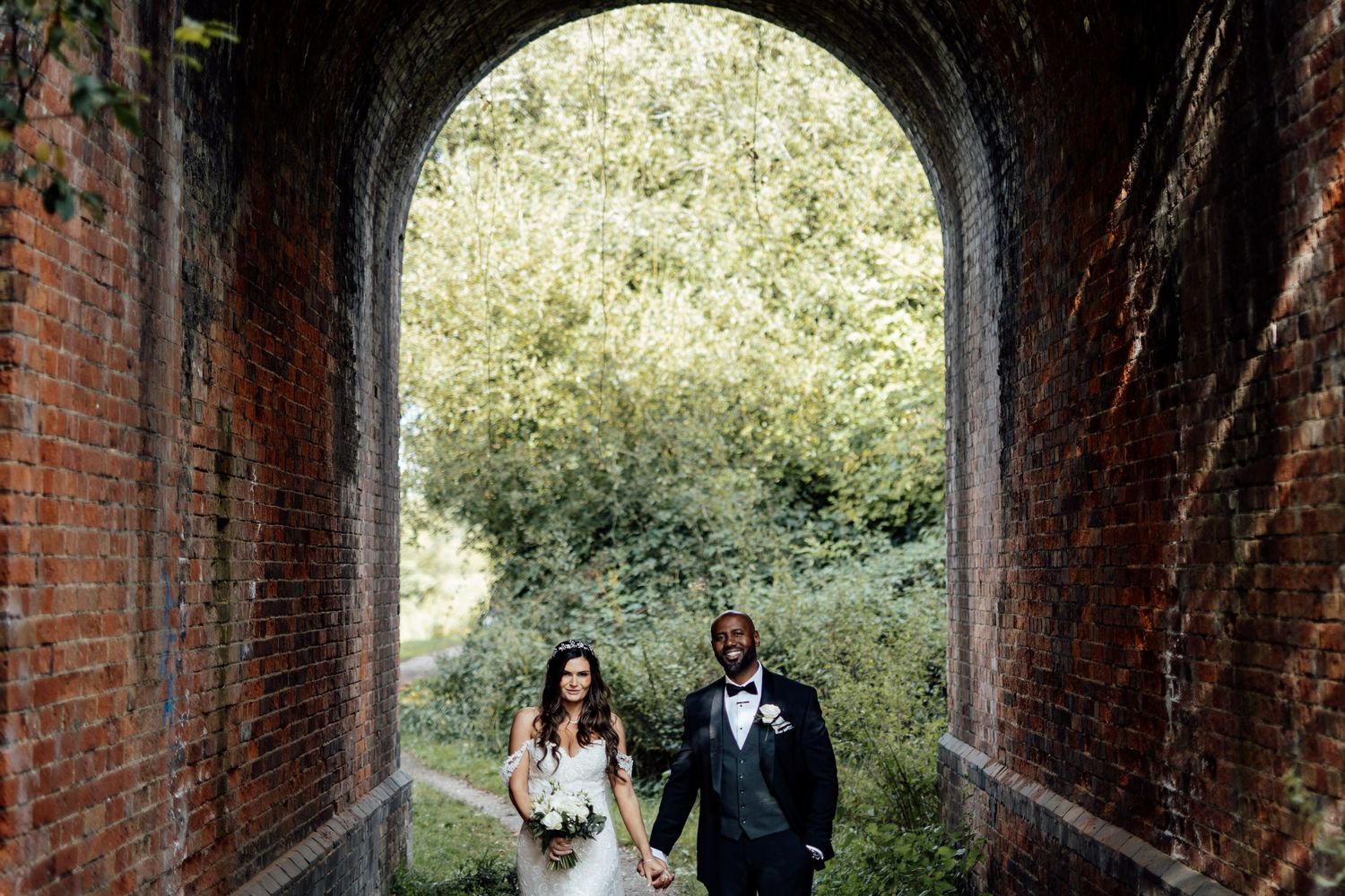 Wedding couple poses in a romantic brick archway surrounded by greenery.