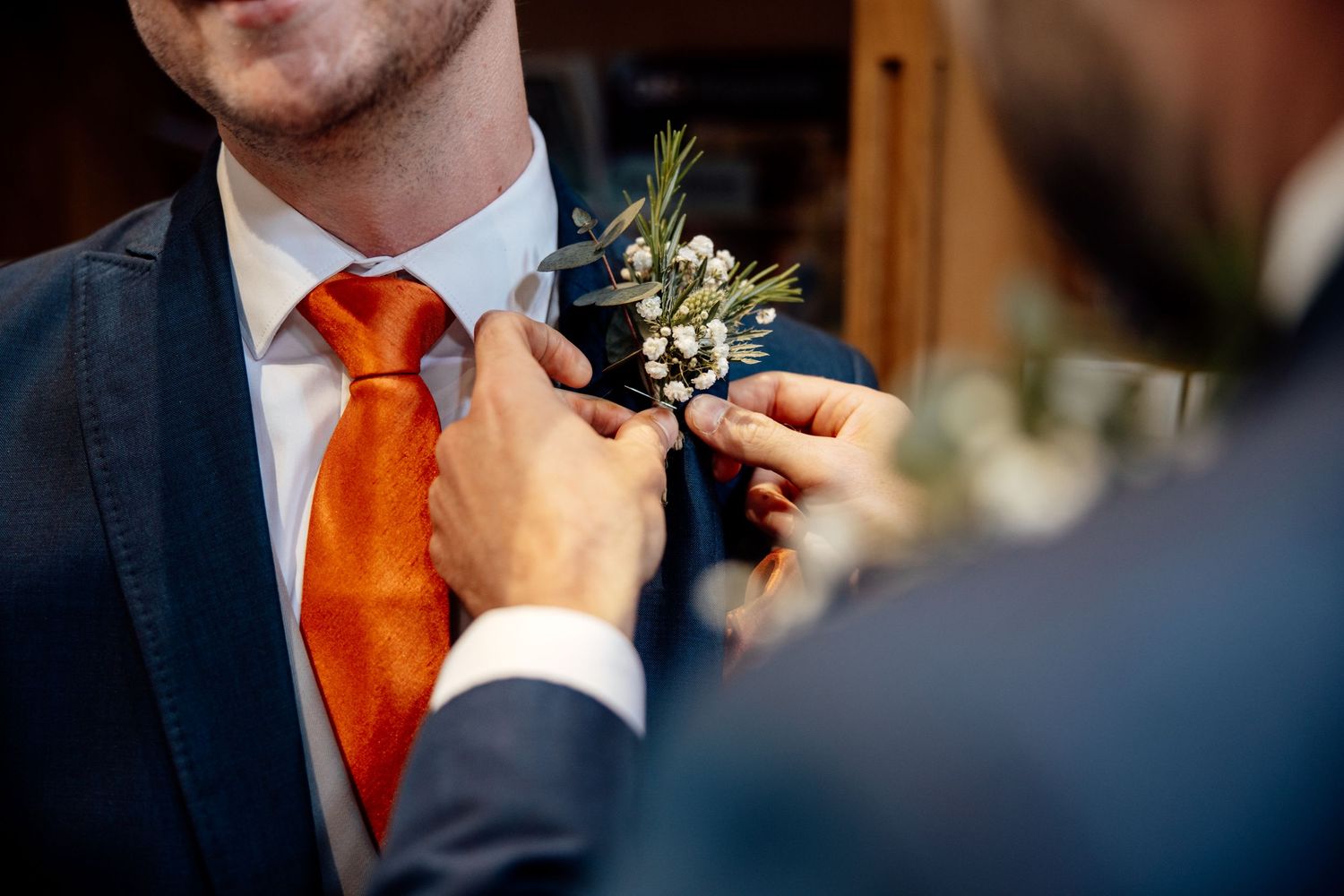 Close-up of boutonniere being pinned to groom's navy suit with orange tie.