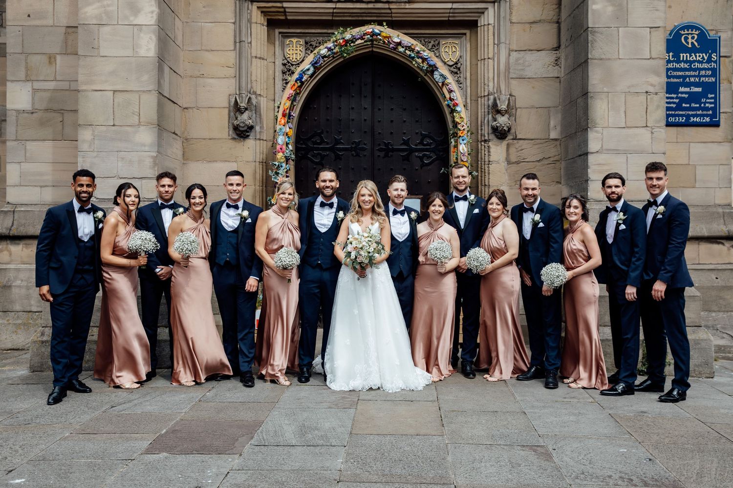 Wedding party poses for formal photo outside historic stone church entrance.