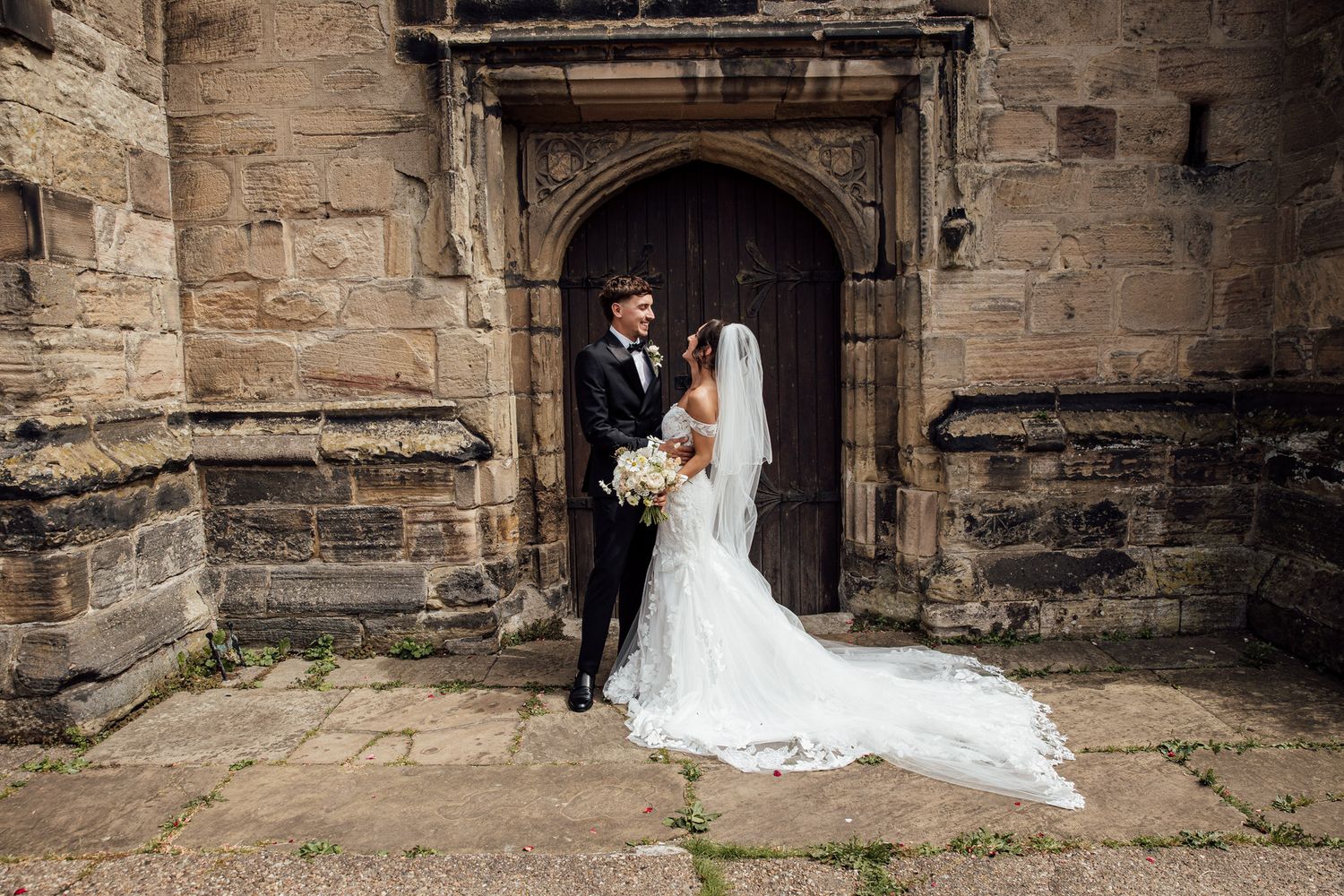Newlyweds pose in front of historic stone archway with flowing white wedding dress train.