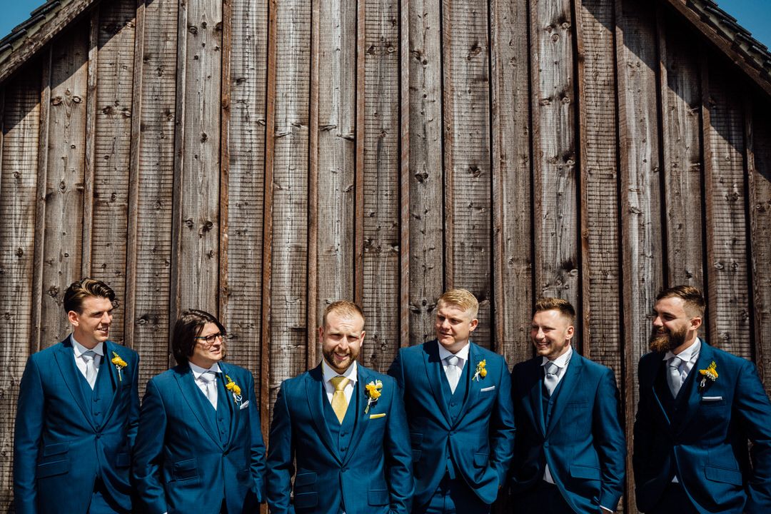 Wedding party wearing coordinated navy blue suits poses against rustic wooden wall backdrop.