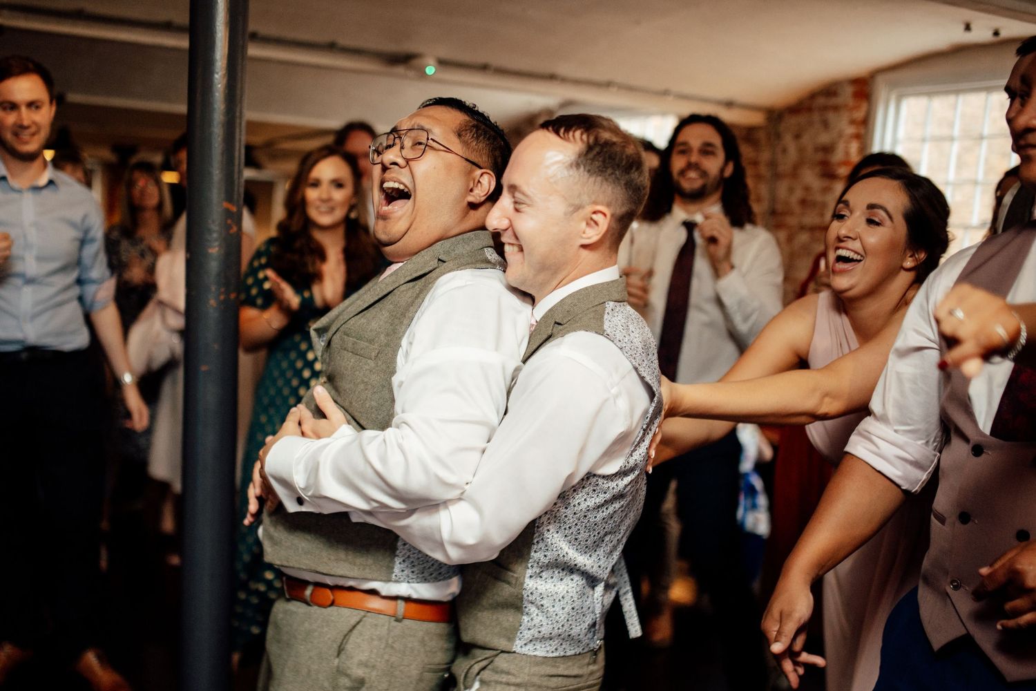 Wedding reception guests dancing and celebrating in a rustic venue with exposed brick walls.