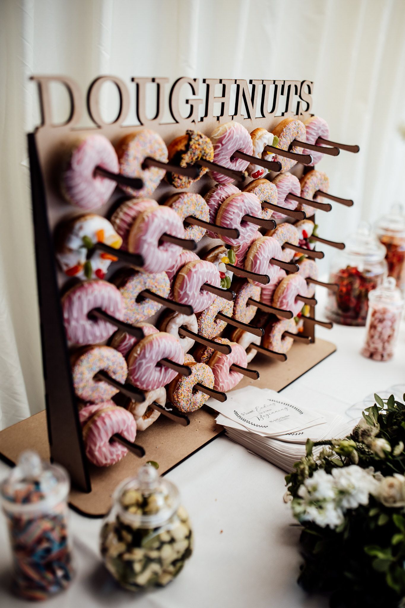 Pink doughnuts displayed on wooden stands with floral decorations at wedding dessert table.