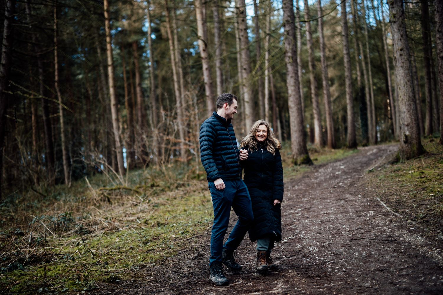 Couple walking together on a winding forest trail surrounded by tall trees.