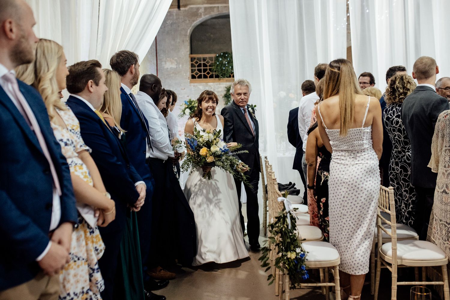 Wedding ceremony in progress with white draped backdrop and floral arrangements.