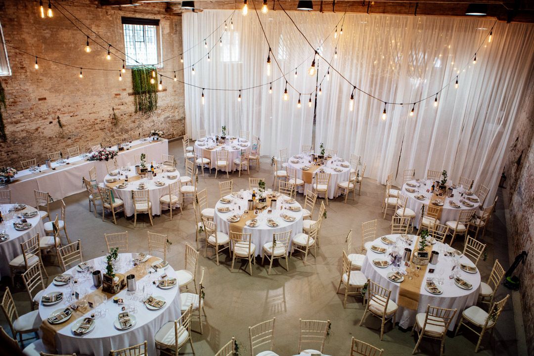 Wedding reception venue with string lights and white draping in rustic industrial space.