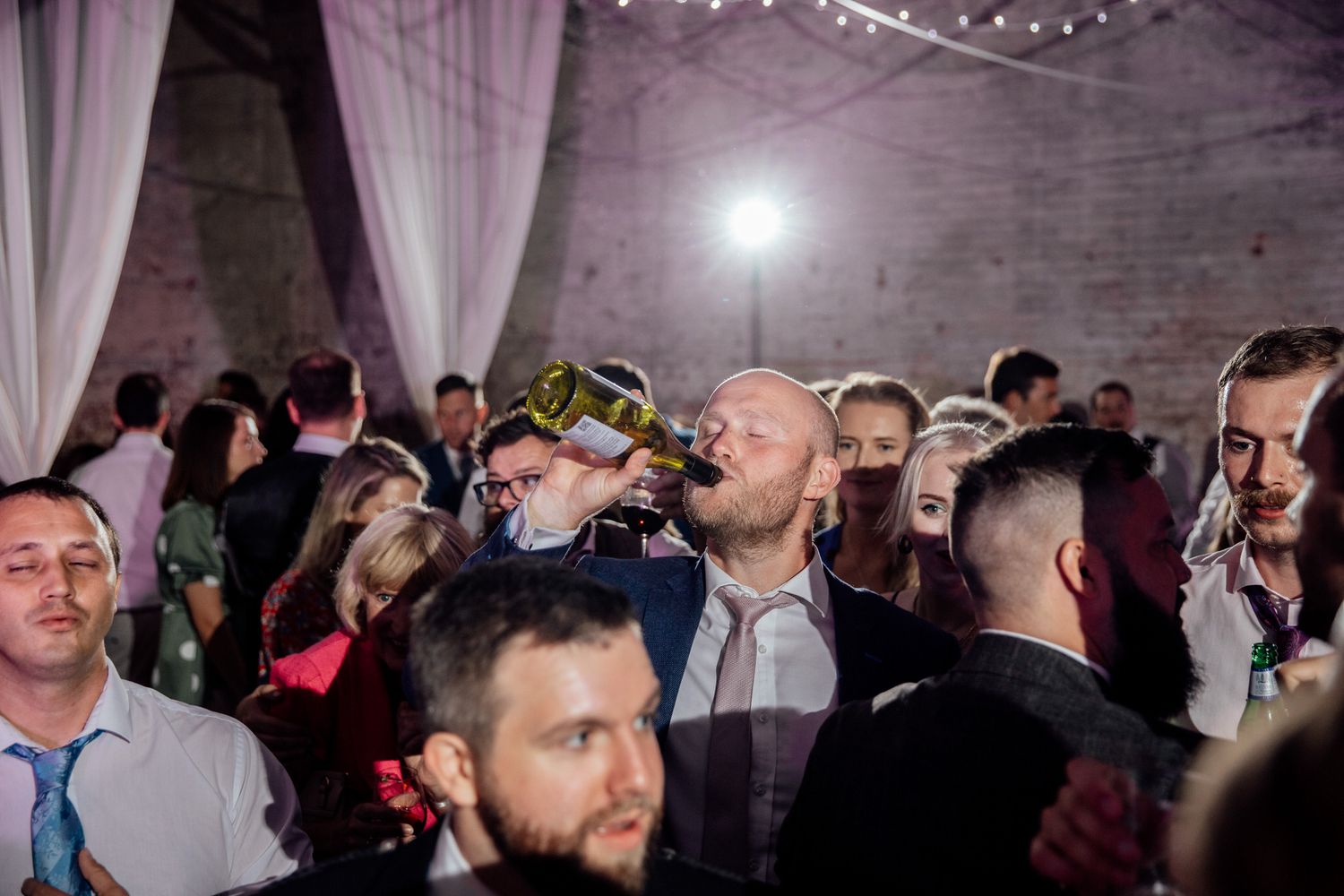 Wedding reception dance floor filled with guests under string lights and purple-tinted lighting.