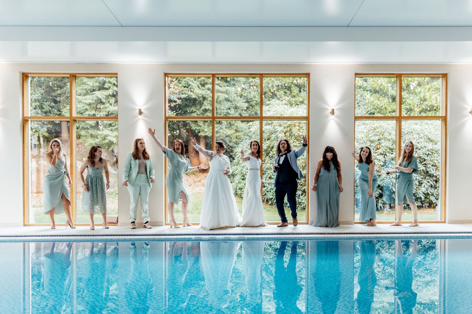 Wedding party poses for photos in front of large windows overlooking nature.