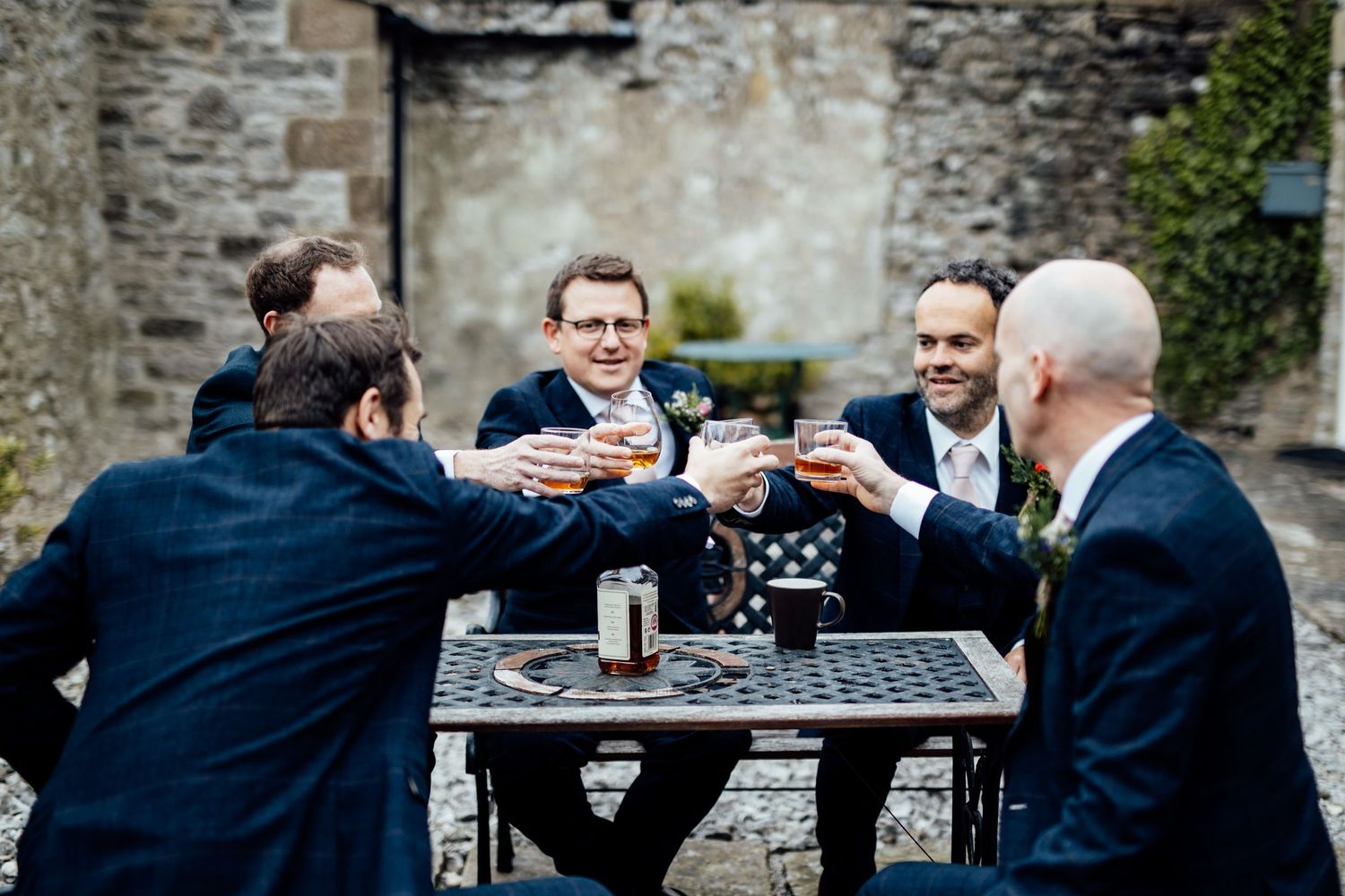 Groomsmen toasting with drinks at an outdoor stone patio table.