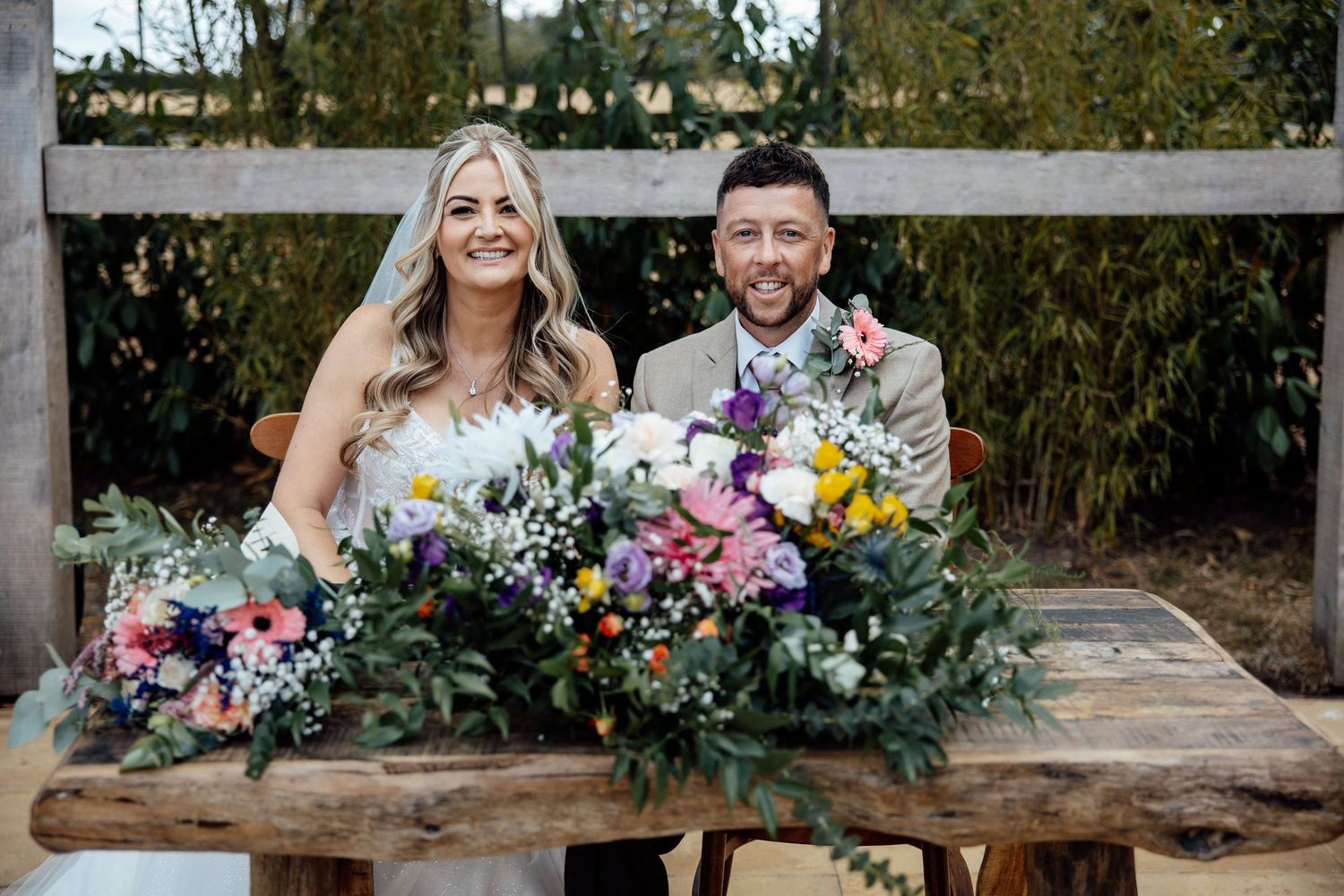 Bride and groom smile at rustic wooden table adorned with colorful wildflower bouquets at outdoor wedding.