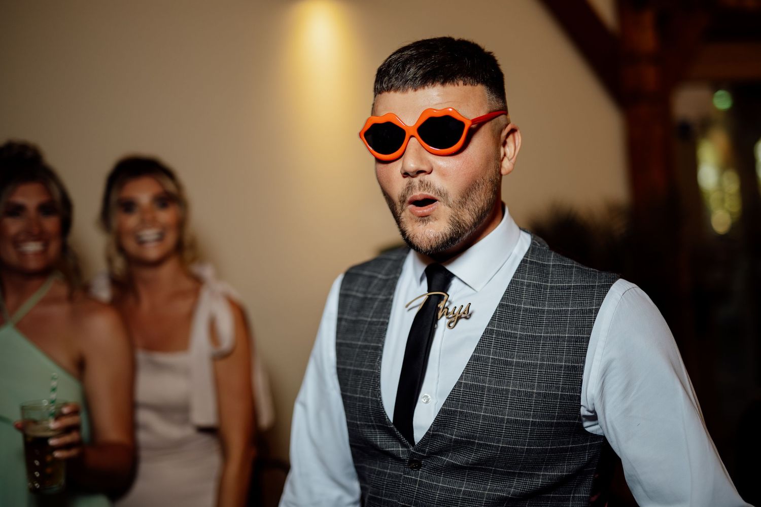 Wedding guest wearing bold orange sunglasses and checked waistcoat poses energetically at reception party.