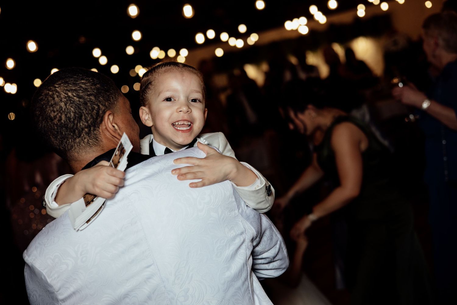Joyful young boy in suit being held by adult dancing under string lights at wedding reception.