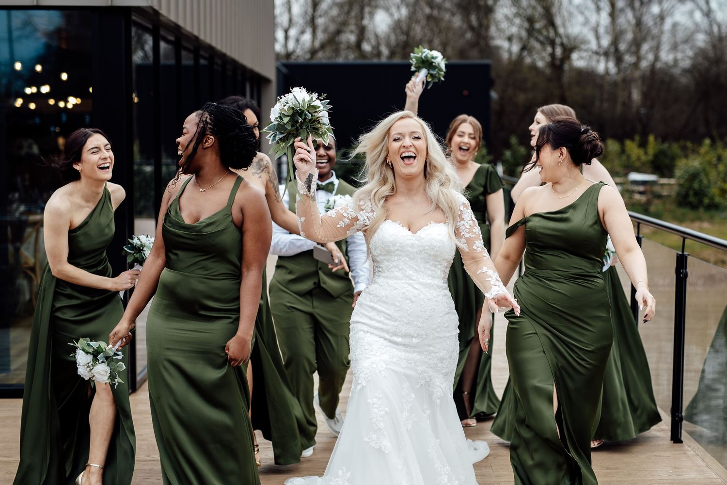 Joyful bride in lace gown celebrating with bridesmaids in olive green dresses outside a modern venue.