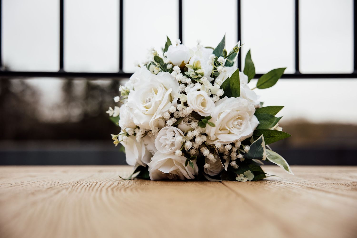 White rose wedding bouquet with greenery and baby's breath on a wooden table.