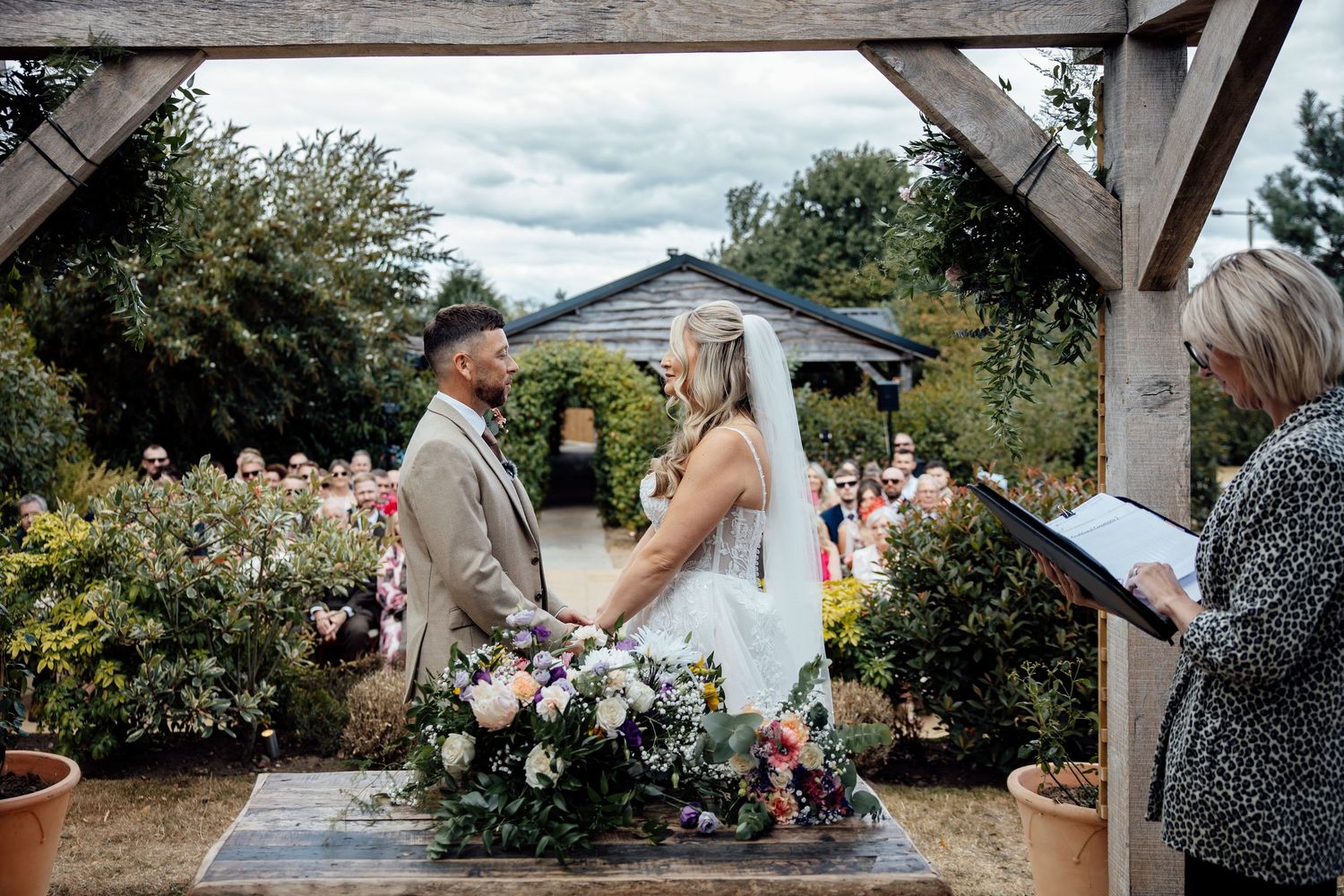 Bride and groom exchange vows outdoors under a wooden arch surrounded by lush gardens and wedding guests.